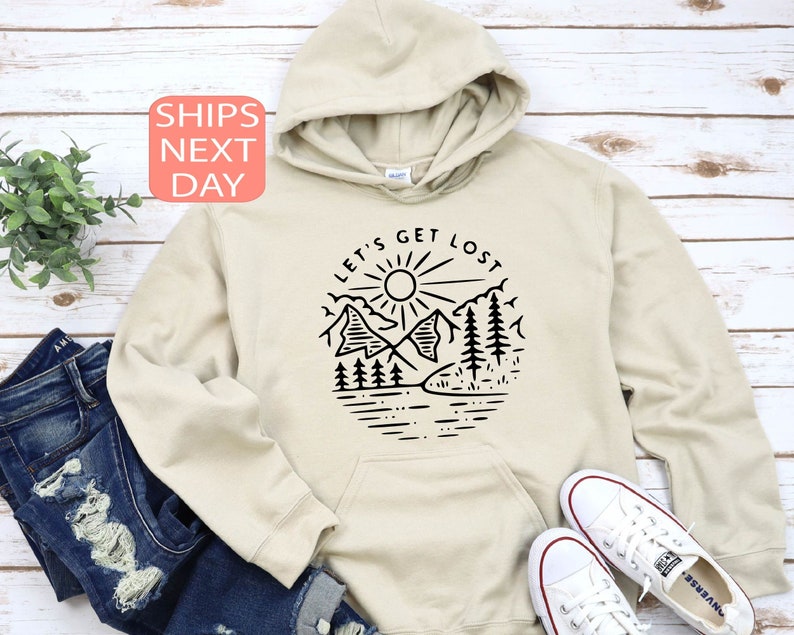May include: A beige hoodie with a black graphic design featuring mountains, trees, a sun, and the text "LET'S GET LOST". The hoodie is paired with blue jeans and white sneakers. The image is set against a light wood background.