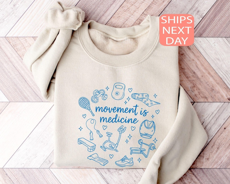 May include: Beige sweatshirt with the phrase "movement is medicine" in blue, surrounded by blue illustrations of fitness equipment. A scrunchie is on the shoulder. The sweatshirt is folded on a wooden surface.