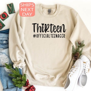 May include: A beige sweatshirt with the text "Thirteen #OfficialTeenager" printed in black. The text is in a handwritten font with a small heart above the word "Thirteen".