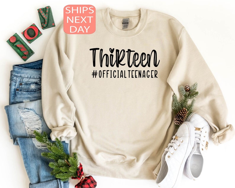 May include: A beige sweatshirt with the text "Thirteen #OfficialTeenager" printed in black. The text is in a handwritten font with a small heart above the word "Thirteen".
