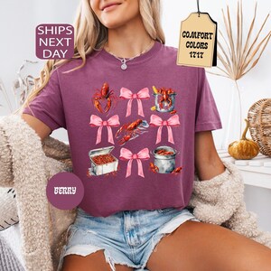 K&ouml;nnte beinhalten: Ein lila T-Shirt mit einem grafischen Aufdruck von Krebsen, einem Topf mit Krebsen und Schleifen. Der Text "COMFORT COLORS 1717" befindet sich auf einem Etikett, das am Shirt befestigt ist.