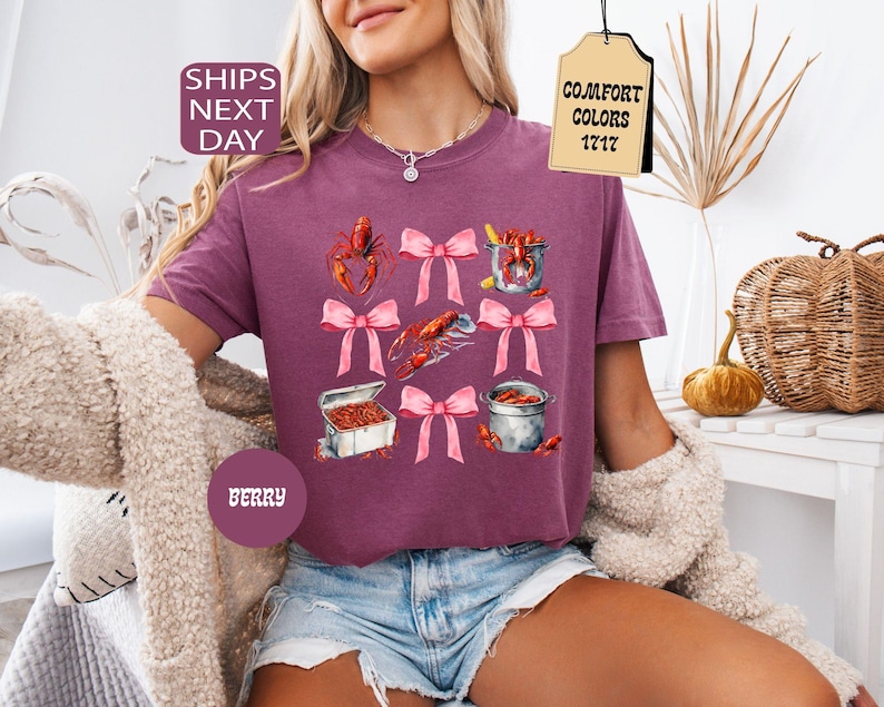 K&ouml;nnte beinhalten: Ein lila T-Shirt mit einem grafischen Aufdruck von Krebsen, einem Topf mit Krebsen und Schleifen. Der Text "COMFORT COLORS 1717" befindet sich auf einem Etikett, das am Shirt befestigt ist.