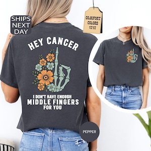 May include: A charcoal grey t-shirt featuring a graphic of a skeleton hand making a middle finger gesture surrounded by orange and light green flowers. The back text reads HEY CANCER I DON'T HAVE ENOUGH MIDDLE FINGERS FOR YOU. A smaller version is on the front.