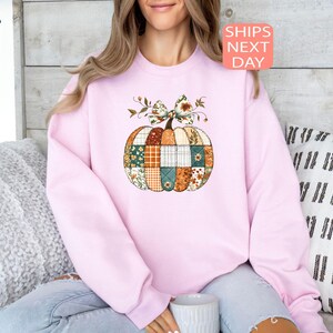 May include: A light pink sweatshirt featuring a patchwork pumpkin design with a floral bow. The pumpkin is composed of various patterned fabrics in shades of orange, green, and blue. The text "SHIPS NEXT DAY" is in a pink box.