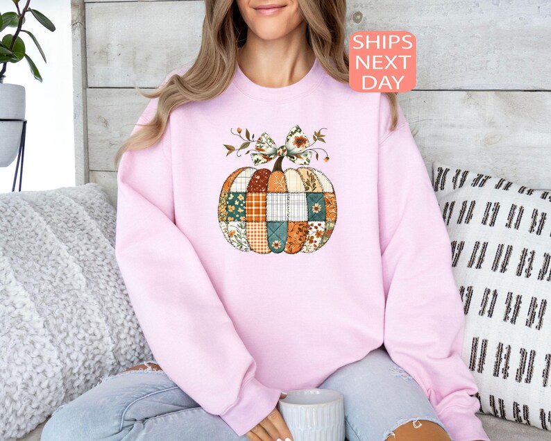 May include: A light pink sweatshirt featuring a patchwork pumpkin design with a floral bow. The pumpkin is composed of various patterned fabrics in shades of orange, green, and blue. The text "SHIPS NEXT DAY" is in a pink box.