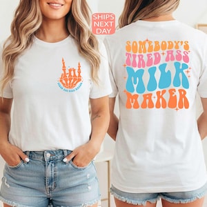 May include: White t-shirt with a skeleton hand graphic making a rock and roll hand gesture. The shirt also has the text "tired ass milk maker" on the front and back in orange, pink, and blue retro font.