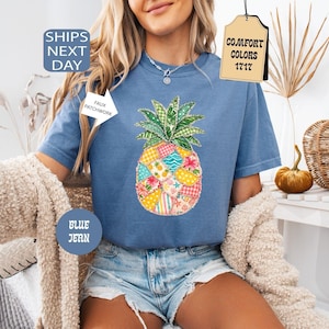 Puede incluir: Una camiseta de manga corta color azul jean con un gráfico grande de piña compuesto por varios patrones de patchwork coloridos, incluyendo diseños florales, de cuadros y de lunares en tonos amarillos, rosas, azules y verdes.