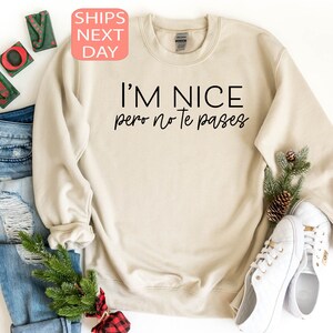 May include: A beige sweatshirt with the text "I'M NICE pero no te pases" in black. The sweatshirt is displayed with a pair of white sneakers, blue jeans, and holiday decorations.