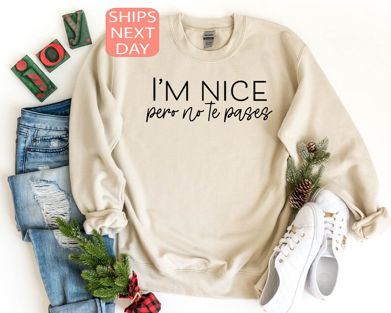 May include: A beige sweatshirt with the text "I'M NICE pero no te pases" in black. The sweatshirt is displayed with a pair of white sneakers, blue jeans, and holiday decorations.