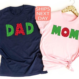 May include: Two t-shirts with watermelon designs. The navy blue shirt says "DAD" and the pink shirt says "MOM".