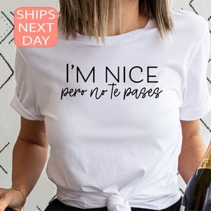 May include: White t-shirt with the text "I'M NICE pero no te pases" in black. The shirt has rolled-up sleeves. A champagne bottle is held in the right hand. The shirt is a casual wear item.