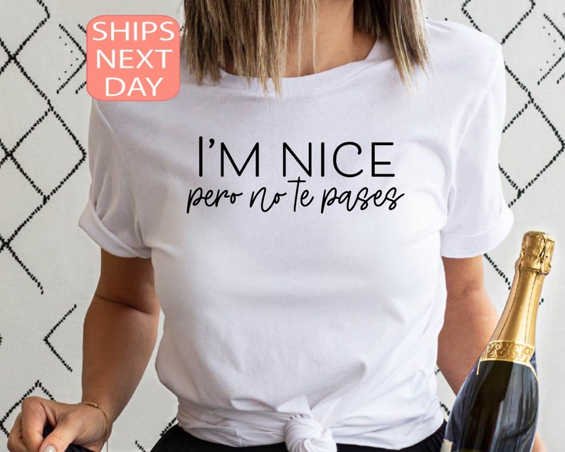 May include: White t-shirt with the text "I'M NICE pero no te pases" in black. The shirt has rolled-up sleeves. A champagne bottle is held in the right hand. The shirt is a casual wear item.