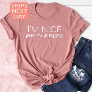 May include: A dusty rose t-shirt with the text "I'M NICE pero no te pases" in white. The shirt is knotted at the waist and paired with blue jeans. A small peach-colored square with the words "SHIPS NEXT DAY" is in the upper left corner.