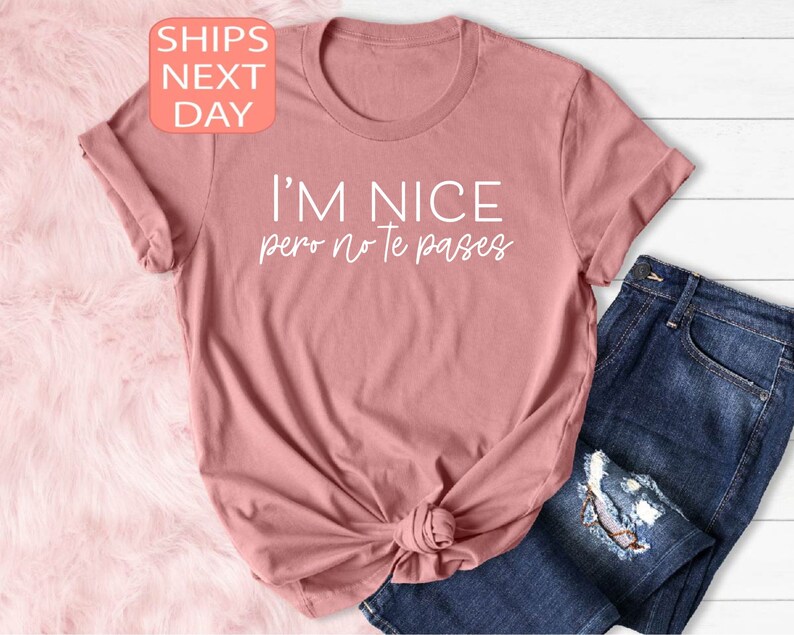 May include: A dusty rose t-shirt with the text "I'M NICE pero no te pases" in white. The shirt is knotted at the waist and paired with blue jeans. A small peach-colored square with the words "SHIPS NEXT DAY" is in the upper left corner.