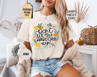 May include: An ivory short-sleeve t-shirt featuring a central graphic with the text "Don't let the hard days win!" surrounded by a wreath of green leaves, orange flowers, and yellow circles. The shirt has a crew neckline and a relaxed fit.