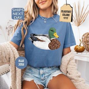 May include: Blue jean t-shirt with a duck applique. The duck features a green plaid head, a yellow beak, and a brown floral patterned body. The shirt has the text "Comfort Colors 1717" on a tag and "Ships Next Day" on a blue circle.