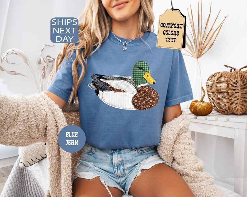 May include: Blue jean t-shirt with a duck applique. The duck features a green plaid head, a yellow beak, and a brown floral patterned body. The shirt has the text "Comfort Colors 1717" on a tag and "Ships Next Day" on a blue circle.