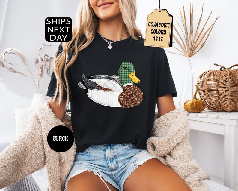 May include: Black t-shirt featuring a patchwork duck design with green, brown, and white patterns. The shirt has the text "Comfort Colors 1717" on a tag and "Ships Next Day" on a black circle.