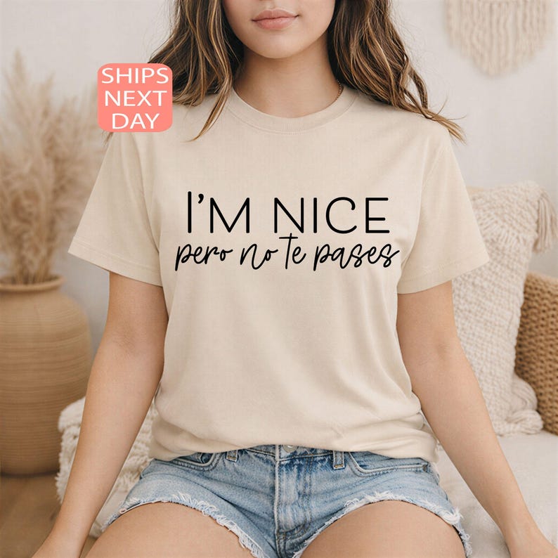 May include: Beige t-shirt with the text "I'M NICE pero no te pases" in black. The shirt has short sleeves and a crew neck. The text is in a bold, sans-serif font. The shirt is paired with denim shorts.
