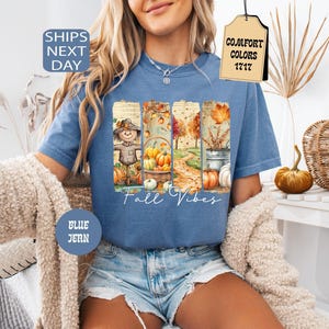 May include: Blue jean t-shirt with a fall-themed graphic. The graphic features a scarecrow, pumpkins, and autumn foliage. The words "Fall Vibes" are written in a script font. The shirt has a round neck and short sleeves.