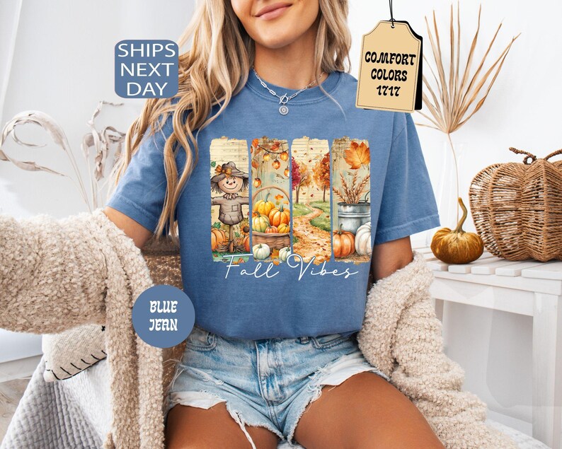 May include: Blue jean t-shirt with a fall-themed graphic. The graphic features a scarecrow, pumpkins, and autumn foliage. The words "Fall Vibes" are written in a script font. The shirt has a round neck and short sleeves.