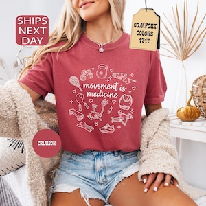 K&ouml;nnte beinhalten: Karmesinrotes T-Shirt mit dem Text "movement is medicine" in einem wei&szlig;en kreisf&ouml;rmigen Design mit Fitnessger&auml;ten. Das Shirt wird mit Jeansshorts und einer cremefarbenen Strickjacke kombiniert.
