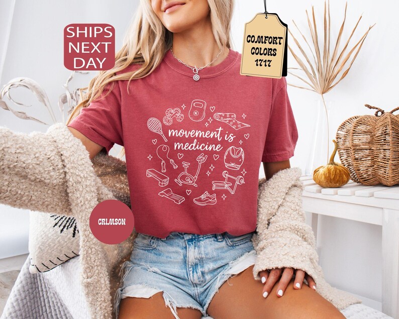 K&ouml;nnte beinhalten: Karmesinrotes T-Shirt mit dem Text "movement is medicine" in einem wei&szlig;en kreisf&ouml;rmigen Design mit Fitnessger&auml;ten. Das Shirt wird mit Jeansshorts und einer cremefarbenen Strickjacke kombiniert.