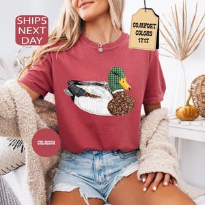 May include: Crimson-colored t-shirt featuring a whimsical duck design with patchwork patterns. The duck has a green plaid head, a brown floral body, and a yellow beak. The shirt has short sleeves and is paired with denim shorts.