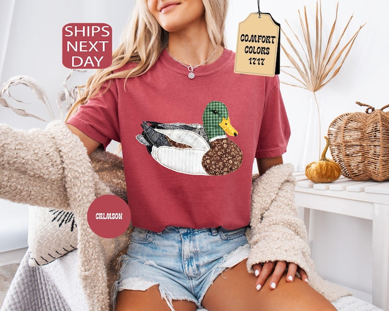 May include: Crimson-colored t-shirt featuring a whimsical duck design with patchwork patterns. The duck has a green plaid head, a brown floral body, and a yellow beak. The shirt has short sleeves and is paired with denim shorts.
