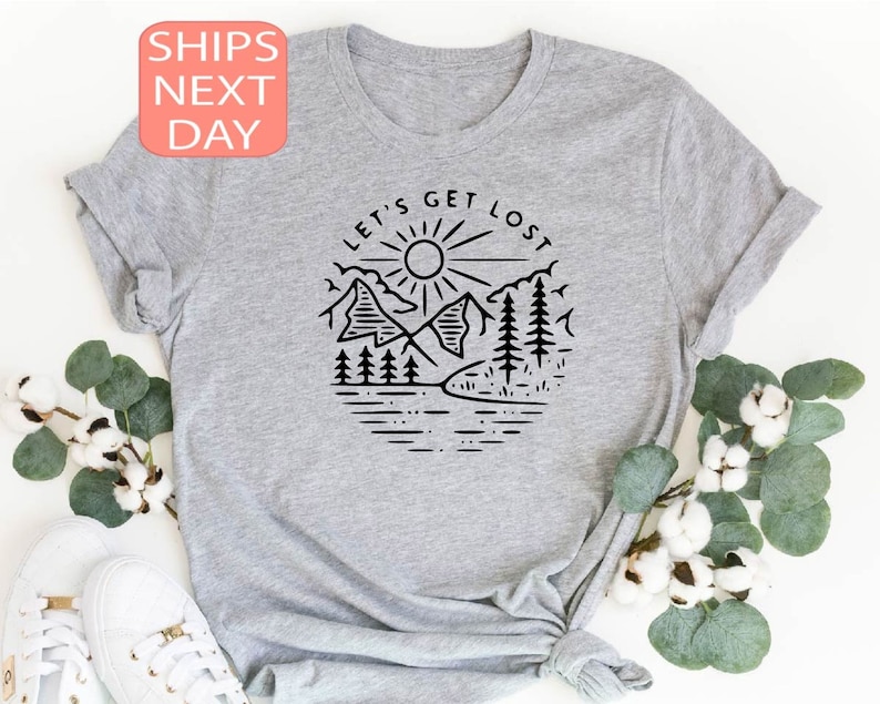 May include: A light gray t-shirt featuring a black graphic of a mountain scene with the text "LET'S GET LOST". The design includes mountains, trees, a sun, and a lake. The shirt is made of a soft material.