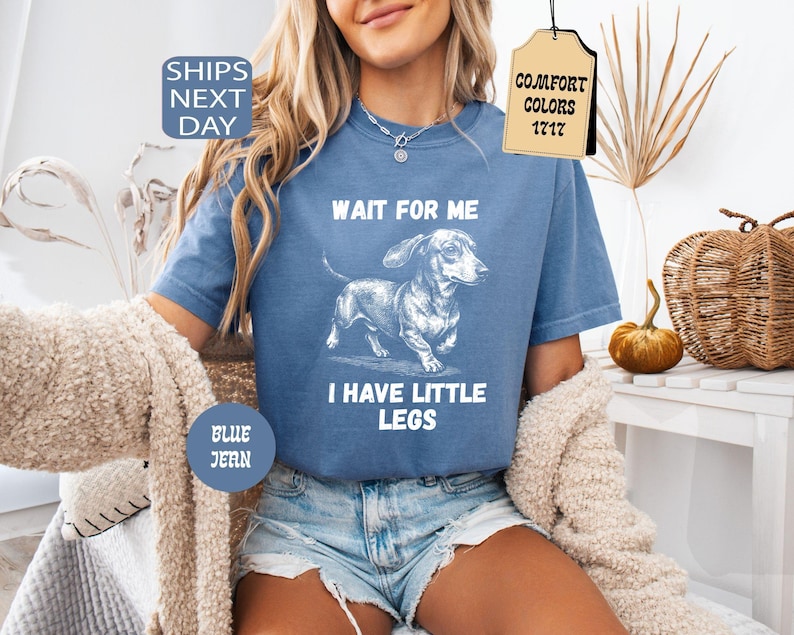 May include: Blue jean-colored t-shirt with a dachshund graphic and the text "WAIT FOR ME I HAVE LITTLE LEGS." The shirt also has the text "COMFORT COLORS 1717" on a tag. The shirt is a comfortable fit.