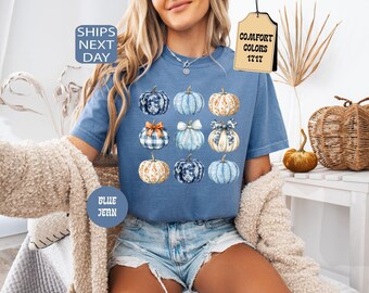 Könnte beinhalten: Ein blaues T-Shirt mit einem Grafikdruck von neun Kürbissen, die in einem Raster angeordnet sind. Die Kürbisse zeigen verschiedene Muster wie Blumen, Karos und einfarbige Designs, einige sind mit dekorativen blauen und orangen Schleifen verziert.