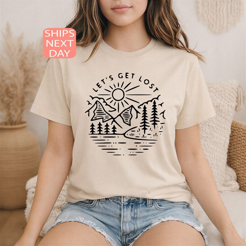 May include: Beige t-shirt with a black circular graphic featuring mountains, trees, a sun, and the text "LET'S GET LOST." The shirt is short-sleeved and has a crew neck. The text "SHIPS NEXT DAY" is in a pink box.