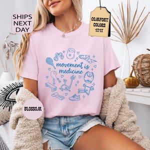 K&ouml;nnte beinhalten: Hellrosa T-Shirt mit dem blauen Schriftzug "movement is medicine", umgeben von Fitness-Illustrationen. Das Shirt hat einen Rundhalsausschnitt und kurze &Auml;rmel. Die W&ouml;rter "Comfort Colors" und "Blossom" sind ebenfalls sichtbar.