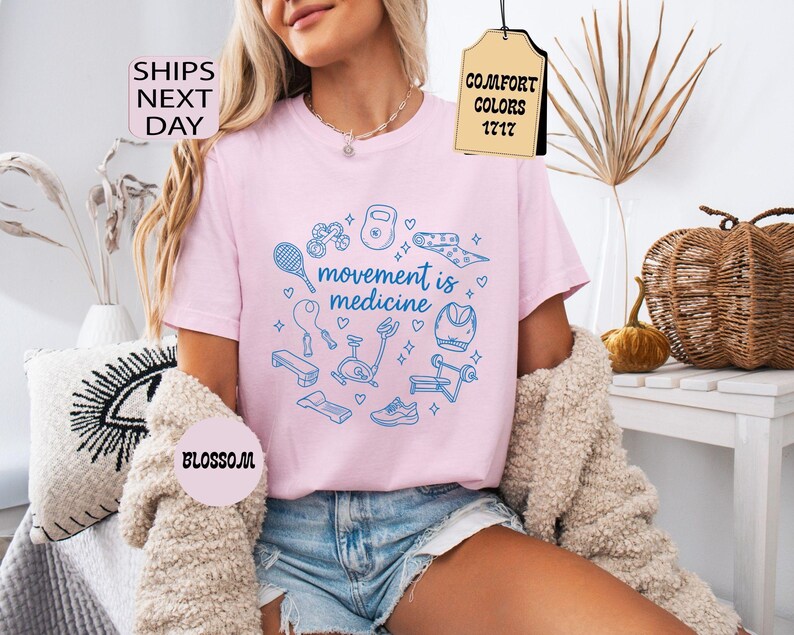 K&ouml;nnte beinhalten: Hellrosa T-Shirt mit dem blauen Schriftzug "movement is medicine", umgeben von Fitness-Illustrationen. Das Shirt hat einen Rundhalsausschnitt und kurze &Auml;rmel. Die W&ouml;rter "Comfort Colors" und "Blossom" sind ebenfalls sichtbar.