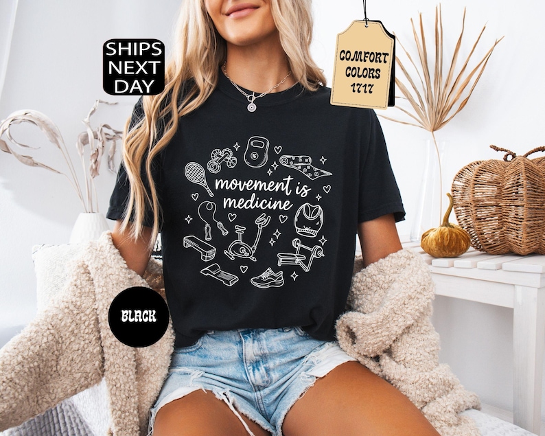 K&ouml;nnte beinhalten: Schwarzes T-Shirt mit wei&szlig;en Strichzeichnungen von Fitnessger&auml;ten und dem Schriftzug "movement is medicine". Das Shirt hat ein Etikett mit der Aufschrift "Comfort Colors 1717". Ein schwarzer Kreis mit dem Wort "BLACK" ist ebenfalls sichtbar.