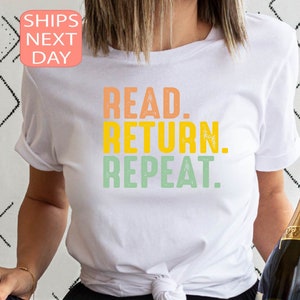 以下が含まれることがあります： オレンジ、黄色、緑で「READ. RETURN. REPEAT.」と書かれた白いTシャツ。袖は折り返されています。背景にはシャンパンのボトルが見えます。