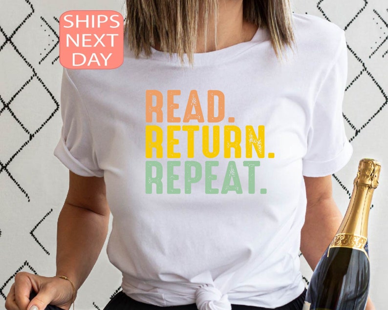 以下が含まれることがあります： オレンジ、黄色、緑で「READ. RETURN. REPEAT.」と書かれた白いTシャツ。袖は折り返されています。背景にはシャンパンのボトルが見えます。