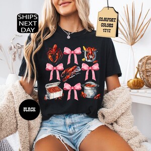 K&ouml;nnte beinhalten: Schwarzes T-Shirt mit einem grafischen Druck von Krebsen, einem Topf mit Krebsen und rosa Schleifen. Der Text "Comfort Colors 1717" befindet sich auf einem braunen Etikett.
