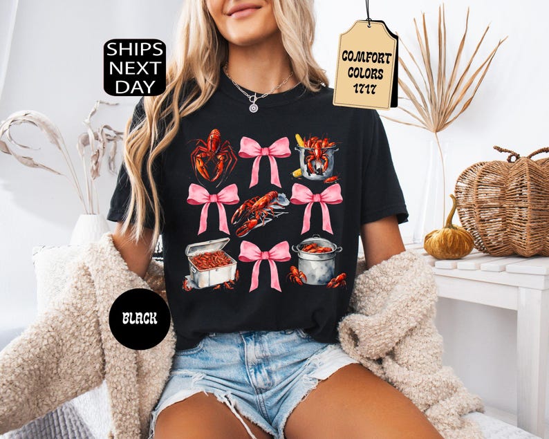 K&ouml;nnte beinhalten: Schwarzes T-Shirt mit einem grafischen Druck von Krebsen, einem Topf mit Krebsen und rosa Schleifen. Der Text "Comfort Colors 1717" befindet sich auf einem braunen Etikett.