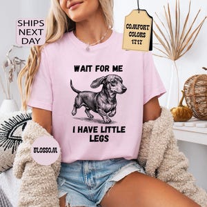 May include: A light pink t-shirt with the text "WAIT FOR ME I HAVE LITTLE LEGS" and a black and white illustration of a dachshund. The shirt has short sleeves and is made of a soft material. The shirt is part of the Comfort Colors 1717 collection.