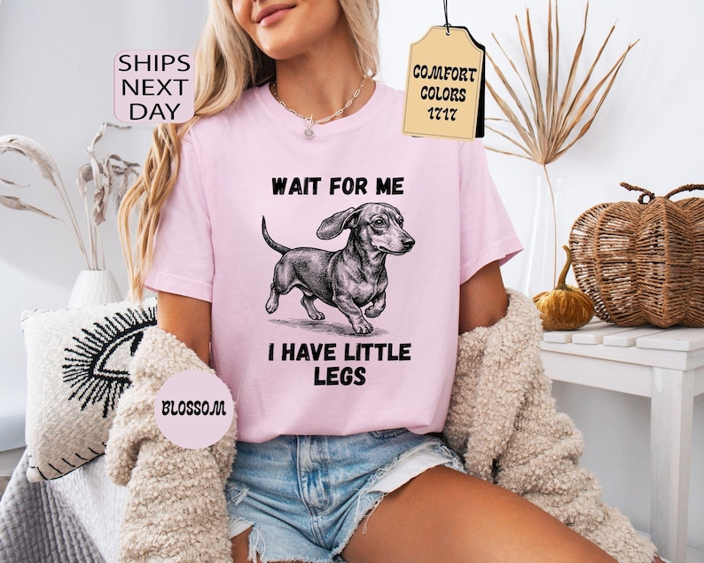 May include: A light pink t-shirt with the text "WAIT FOR ME I HAVE LITTLE LEGS" and a black and white illustration of a dachshund. The shirt has short sleeves and is made of a soft material. The shirt is part of the Comfort Colors 1717 collection.