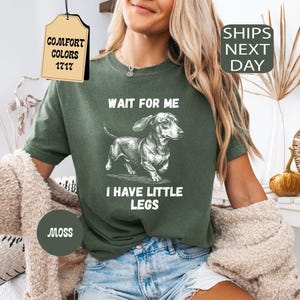 May include: Moss green t-shirt with a dachshund graphic and the text "WAIT FOR ME I HAVE LITTLE LEGS." The shirt also has a tag that says "COMFORT COLORS 1717." The shirt is paired with blue denim shorts.