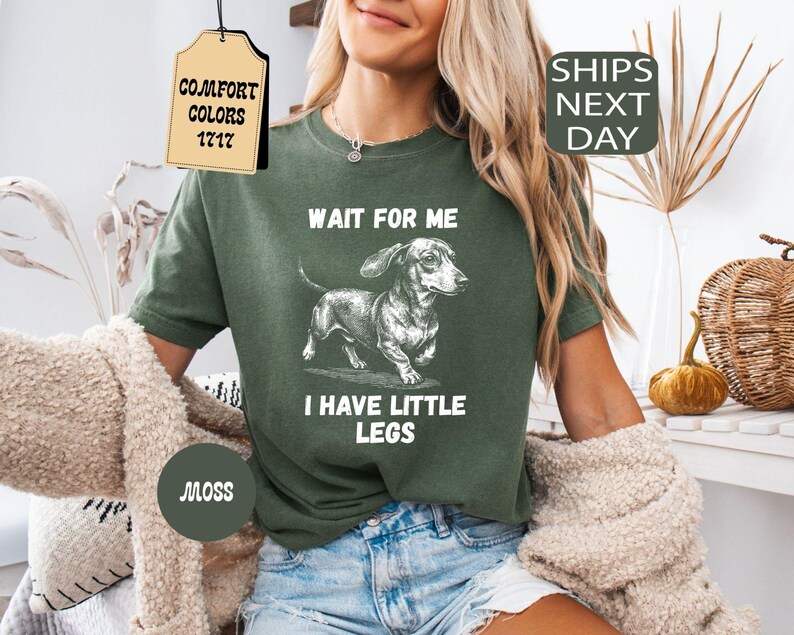 May include: Moss green t-shirt with a dachshund graphic and the text "WAIT FOR ME I HAVE LITTLE LEGS." The shirt also has a tag that says "COMFORT COLORS 1717." The shirt is paired with blue denim shorts.