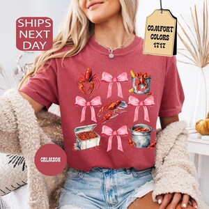 K&ouml;nnte beinhalten: Ein T-Shirt in kirschroter Farbe mit einem grafischen Aufdruck von Krebsen, rosafarbenen Schleifen und dem Text "COMFORT COLORS 1717".