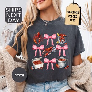 K&ouml;nnte beinhalten: Ein dunkelgraues T-Shirt mit einem grafischen Druck von rosa Schleifen und Krebsen. Das Shirt hat den Text "COMFORT COLORS 1717" auf einem Etikett.