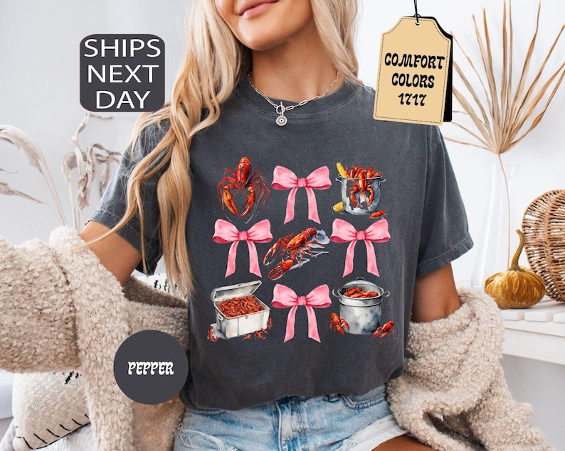 K&ouml;nnte beinhalten: Ein dunkelgraues T-Shirt mit einem grafischen Druck von rosa Schleifen und Krebsen. Das Shirt hat den Text "COMFORT COLORS 1717" auf einem Etikett.