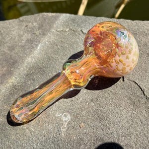 May include: A hand-blown glass pipe with a rounded bowl and a long stem. The pipe features a blend of orange, yellow, and iridescent colors. The glass has a textured appearance, and the pipe is resting on a gray stone surface.