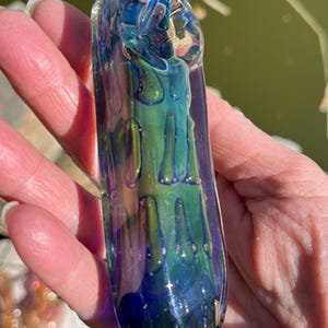 May include: A cylindrical glass pipe featuring a deep blue and iridescent green color palette with a raised, textured drip-like pattern across the surface. The glass has a glossy finish and displays shifting metallic hues under the light.