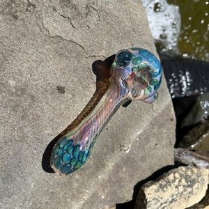 Könnte beinhalten: Eine farbenfrohe Glaspfeife mit Kopf und Stiel. Der Kopf ist mit teal- und grünen blasenartigen Designs verziert, während der Stiel ein wirbelndes Muster aus Rosa, Lila und Blau aufweist. Die Pfeife liegt auf einer grauen Steinoberfläche.